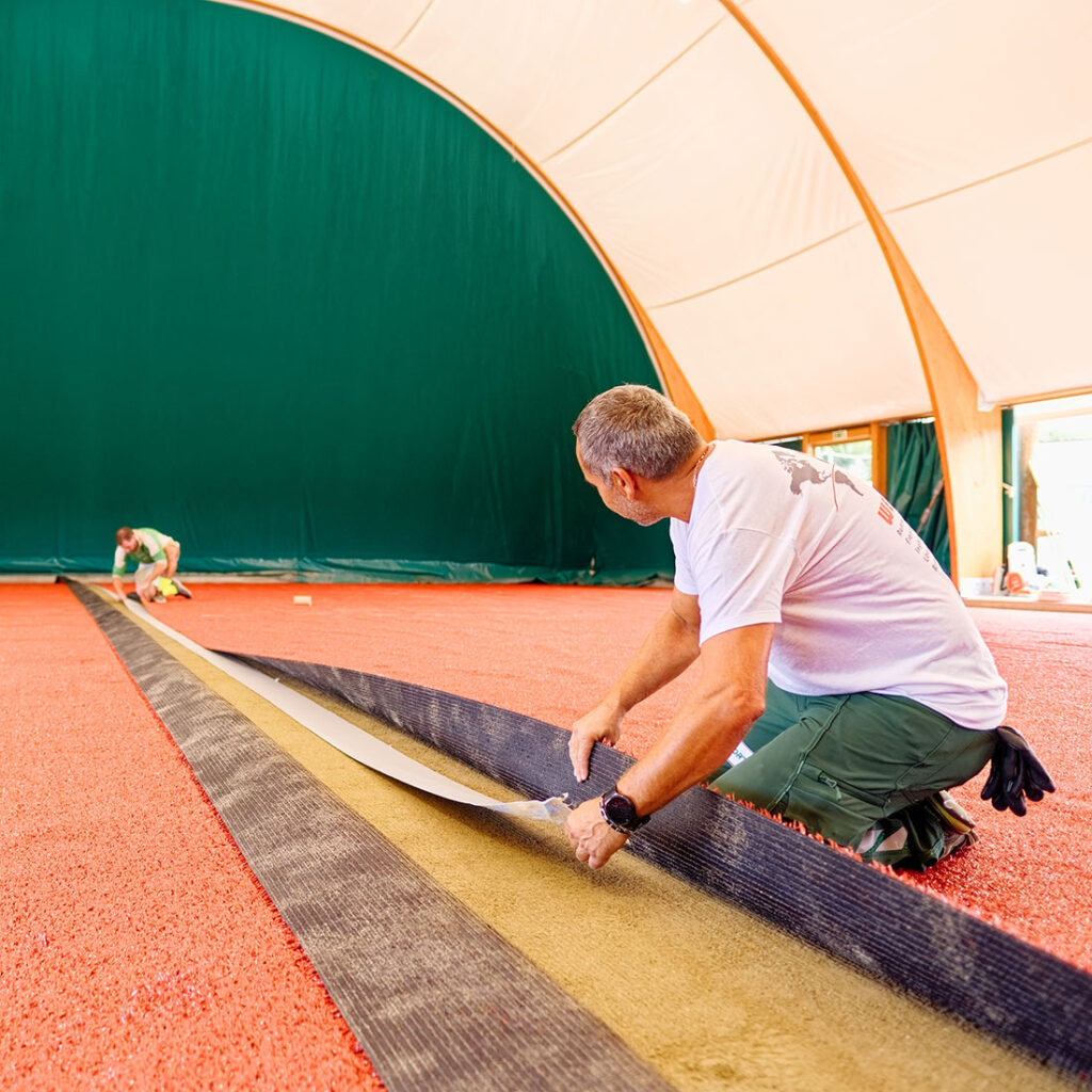 Medewerker van RedPlus aan het werk aan de naden bij het onderhoud van een rode gravelbaan in een hal